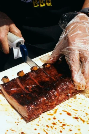 Close-up of person in plastic gloves slicing saucy ribs on a white cutting board; BBQ text partially visible in background.