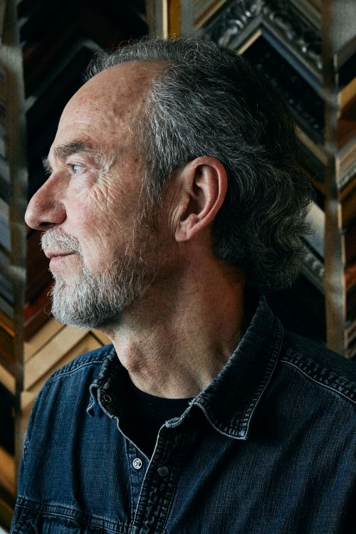 Portrait of an older adult male in a denim shirt. He has graying hair and is set against a background of picture frame samples.