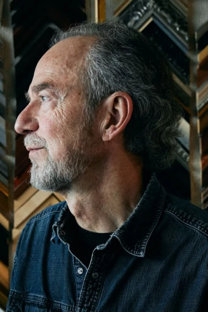 Portrait of an older adult male in a denim shirt. He has graying hair and is set against a background of picture frame samples.