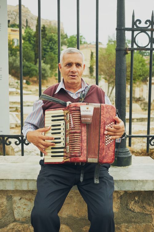 Elderly man plays a red accordion while seated on a stone wall in an urban setting with old buildings and trees in the background.