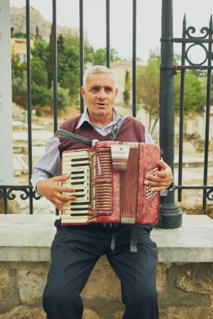 Elderly man plays a red accordion while seated on a stone wall in an urban setting with old buildings and trees in the background.