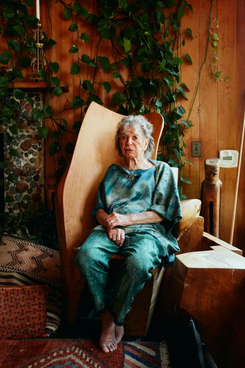 Elderly woman with short grey hair sitting in a wooden chair indoors, wearing blue tie-dye clothing, with plants and wood paneling.
