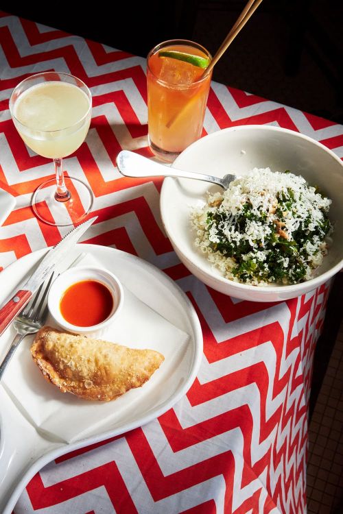 Overhead shot of a table setting with two cocktails, a bowl of salad, a pastry with red sauce, and cutlery on a patterned tablecloth.
