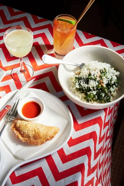 Overhead shot of a table setting with two cocktails, a bowl of salad, a pastry with red sauce, and cutlery on a patterned tablecloth.