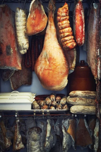 Selection of cured meats and sausages hanging and displayed on shelves, variety of colors and textures in a food storage setting.