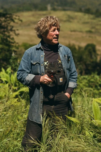 An adult male in denim jacket and overalls holds a wine glass outdoors in a sunny field, enjoying a glass of red wine.