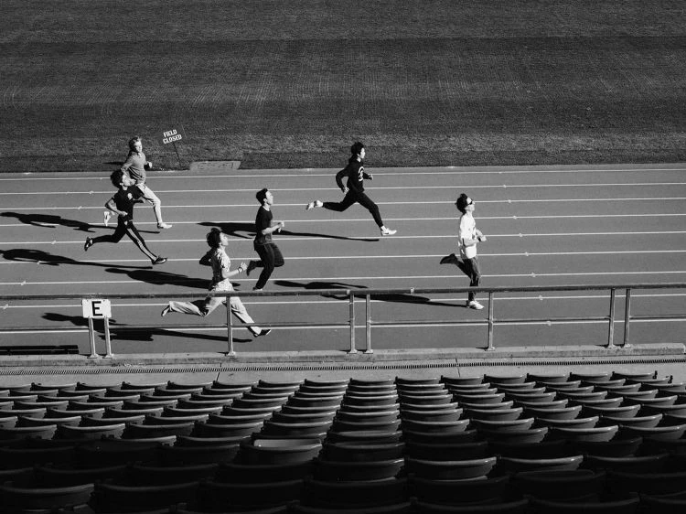 Monochrome, high-angle shot of six runners racing on an outdoor track with stadium seating visible below.