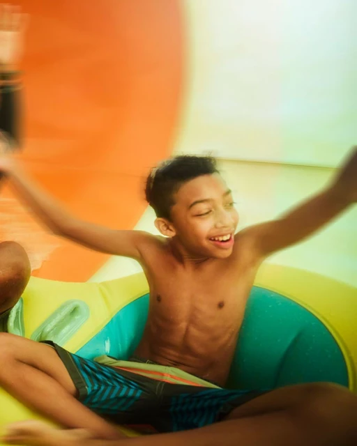 Smiling child enjoys an inflatable pool with hands raised, wearing blue patterned shorts, creating a vivid, joyful summer image.