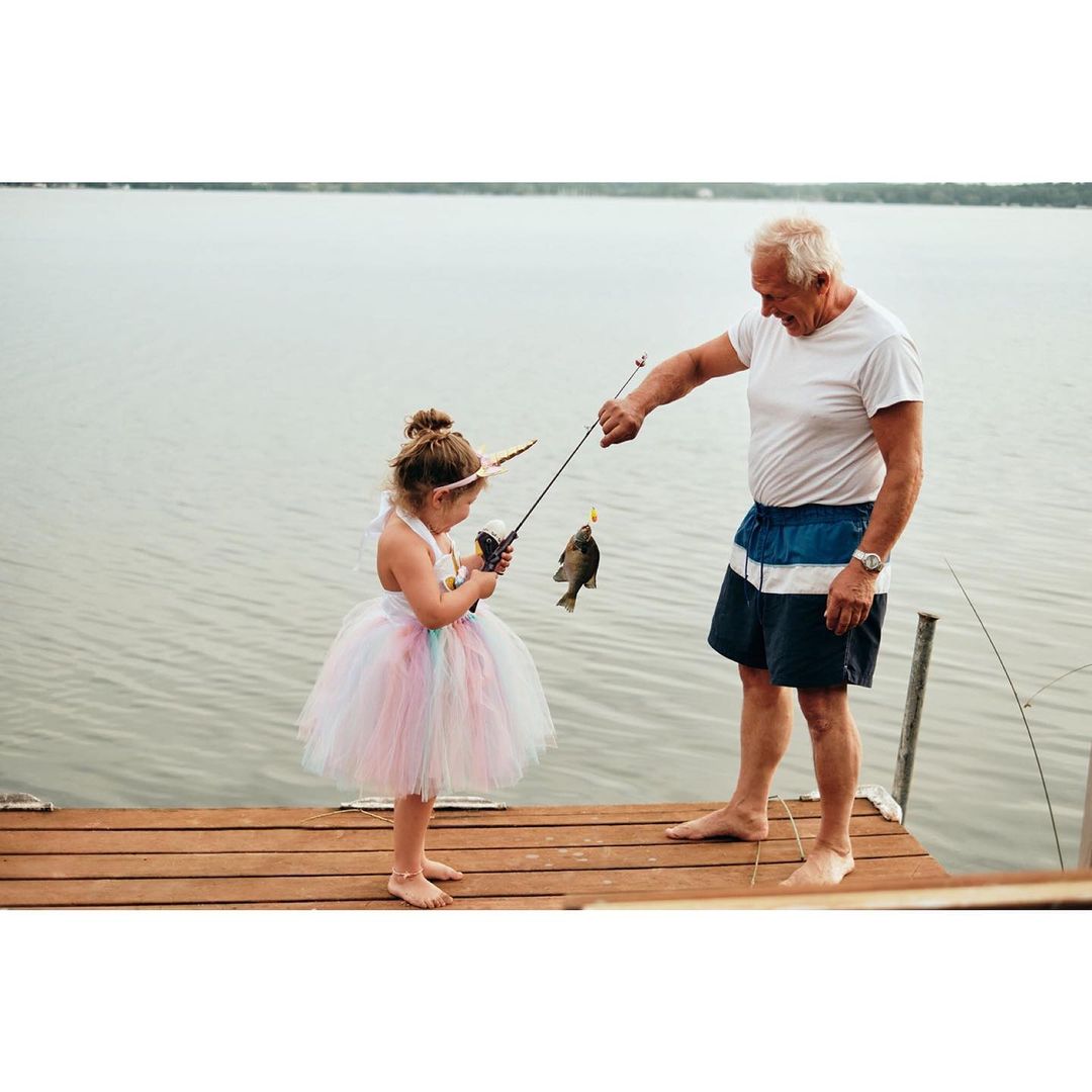 A little girl in a tutu and an older man hold a fishing rod with a fish on a pier near a lake, enjoying a shared outdoor activity.