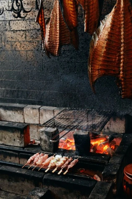 Close-up shot of skewers with meat grilling over hot coals indoors, with brown, smoked fish hanging above a brick oven.