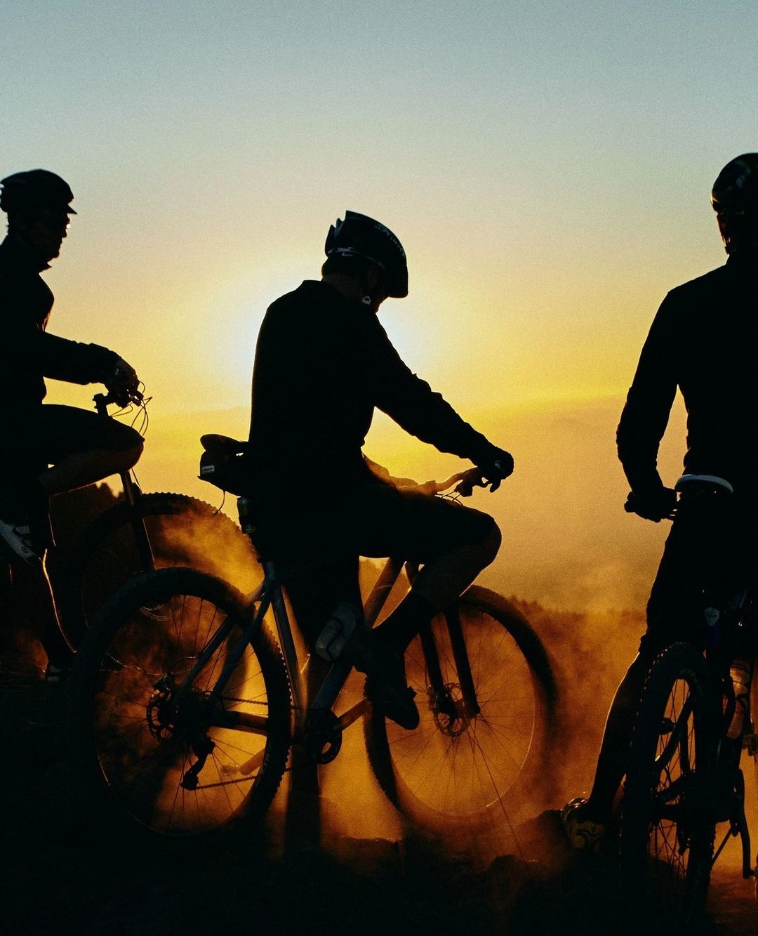 Three cyclists in silhouette riding mountain bikes against a bright sunset sky, kicking up dust from the ground.