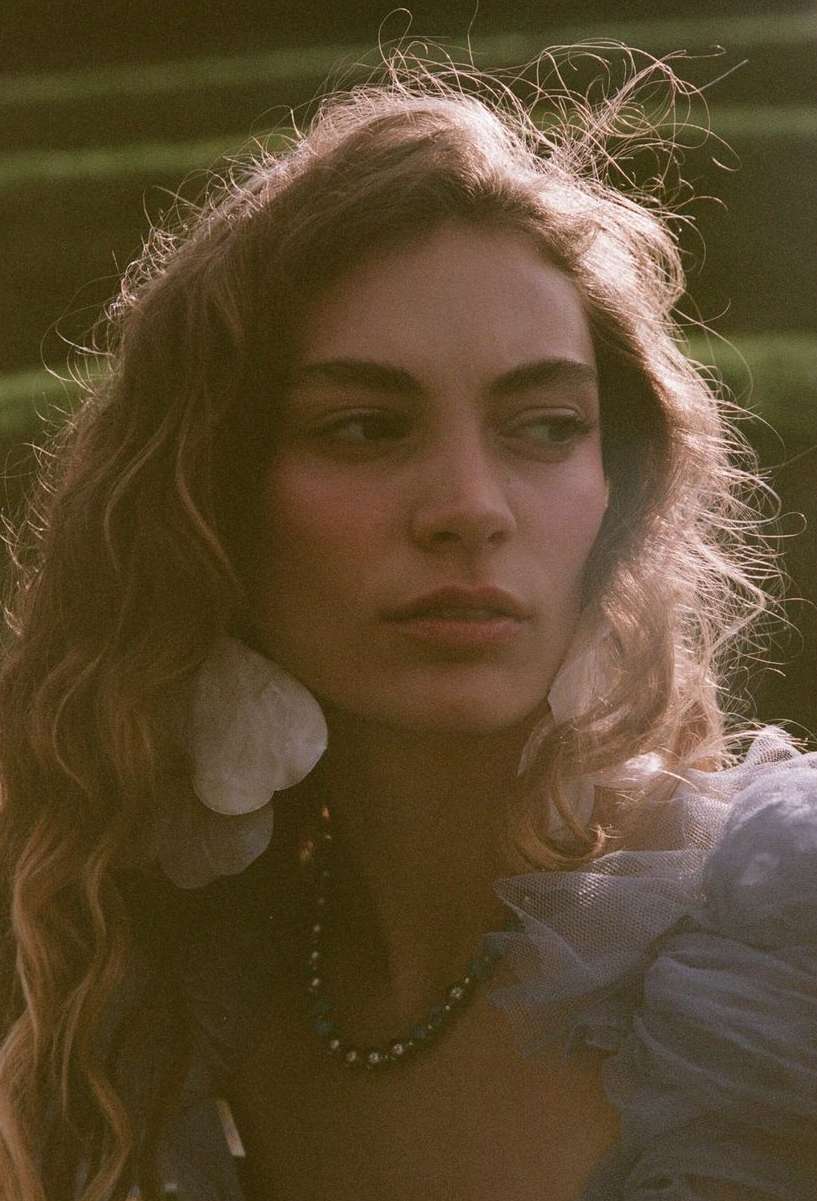 Close-up portrait of an adult woman with wavy hair, wearing large floral earrings and a beaded necklace, against a green background.