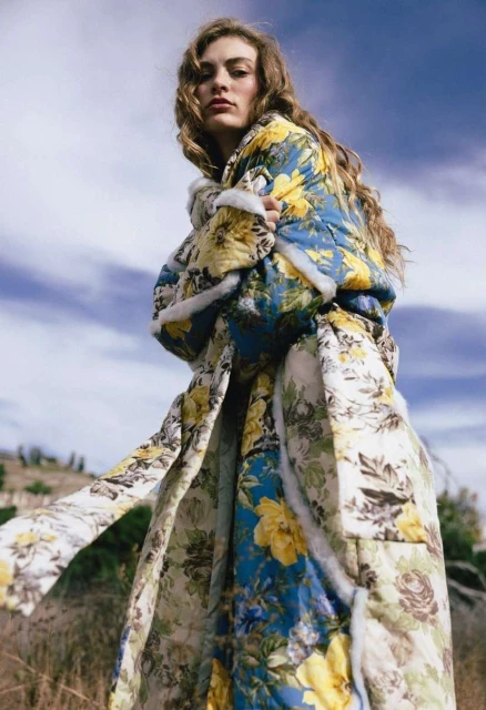 Woman wearing a floral-patterned coat standing in a field with a blue sky and clouds in the background, fashion editorial shot.