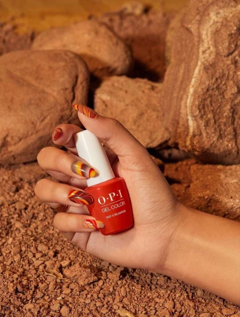 Close-up of a woman's hand holding a bottle of red OPI gel nail polish against a backdrop of brown rocks and earth tones. Focus on product.