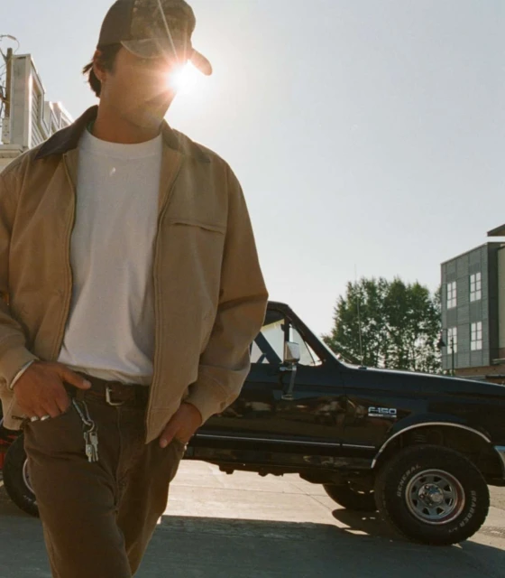 Man in jacket and baseball cap standing next to an old pickup truck on a bright sunny day in an outdoor urban environment.
