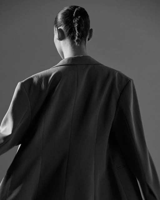 Back view of an adult woman with a braided hairstyle wearing a blazer against a grey background in a studio setting.