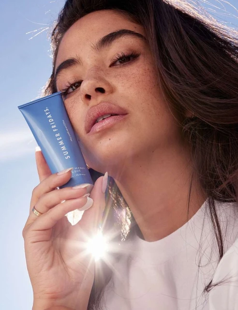 Woman holds Summer Fridays skincare tube against a blue sky backdrop, product promotion for beauty and personal care.