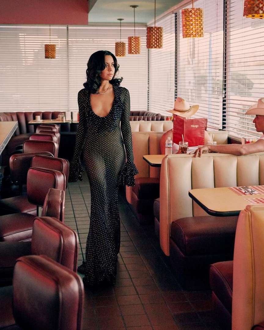 Elegant woman in a black polka-dot dress walks through a vintage diner, with booths and retro pendant lights enhancing the classic setting.