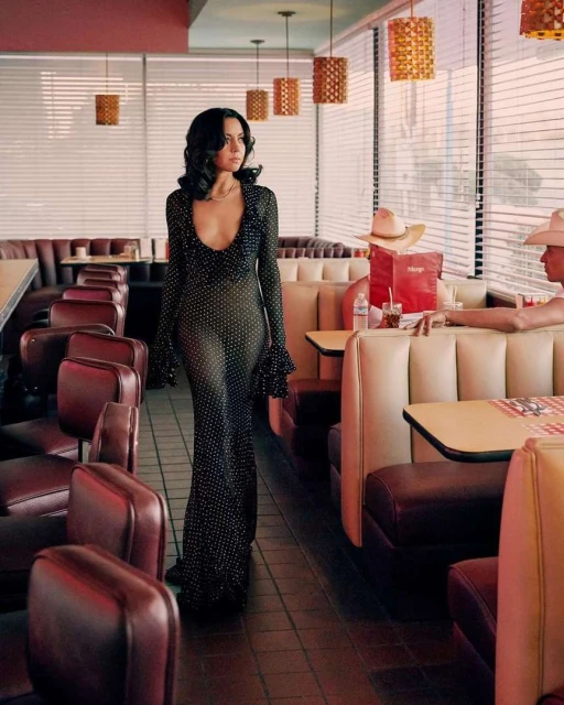 Elegant woman in a black polka-dot dress walks through a vintage diner, with booths and retro pendant lights enhancing the classic setting.