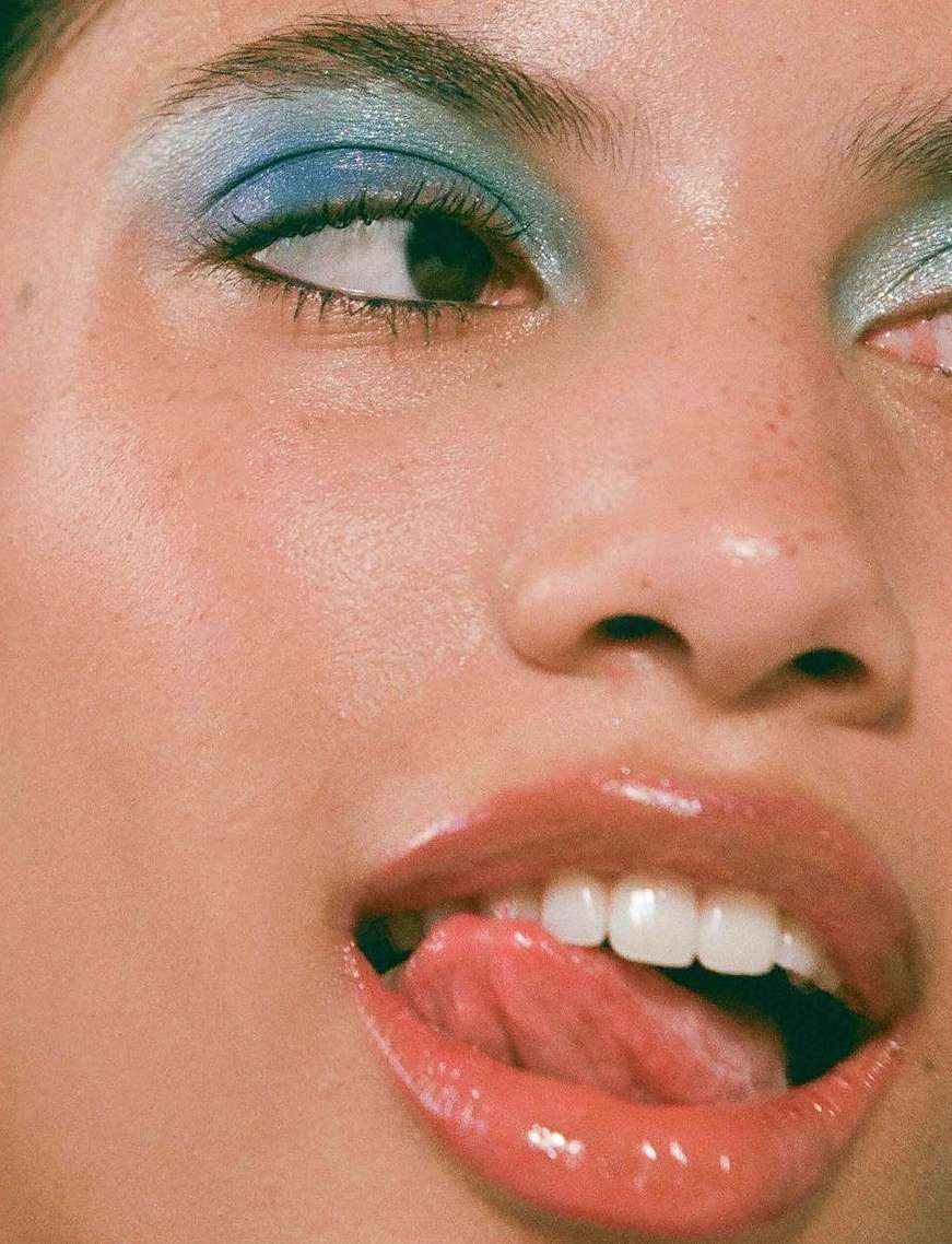 Close-up of a woman's face with blue eyeshadow and glossy lips, sticking her tongue out, showcasing makeup and beauty.