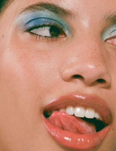 Close-up of a woman's face with blue eyeshadow and glossy lips, sticking her tongue out, showcasing makeup and beauty.