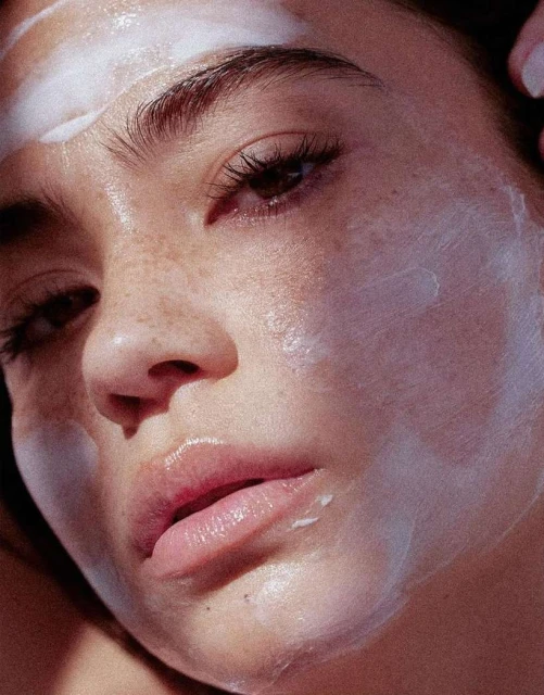 Close-up of a young adult woman with a white facial mask, soft lighting, and visible freckles in a beauty or skincare context.