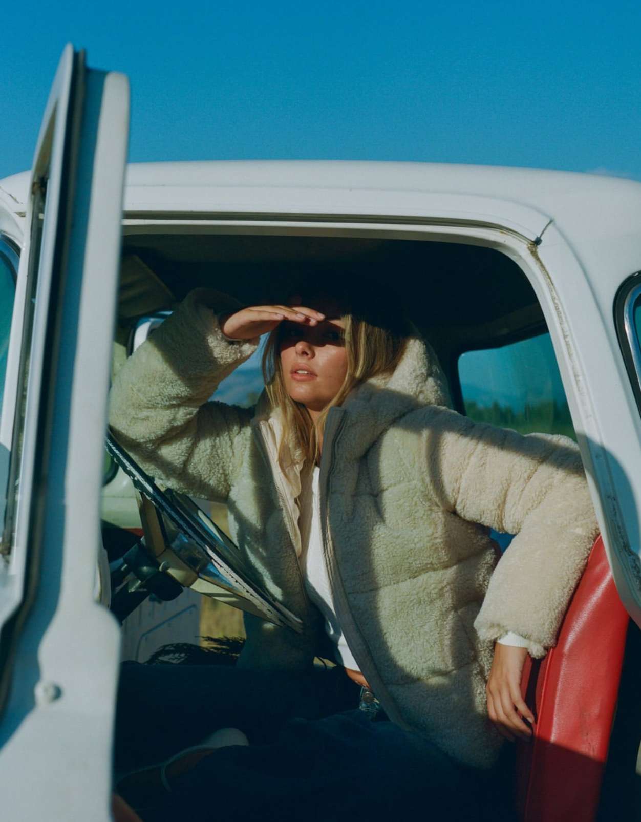 A woman shields her eyes while looking out from inside a vintage car, wearing a fluffy jacket against a clear blue sky backdrop.