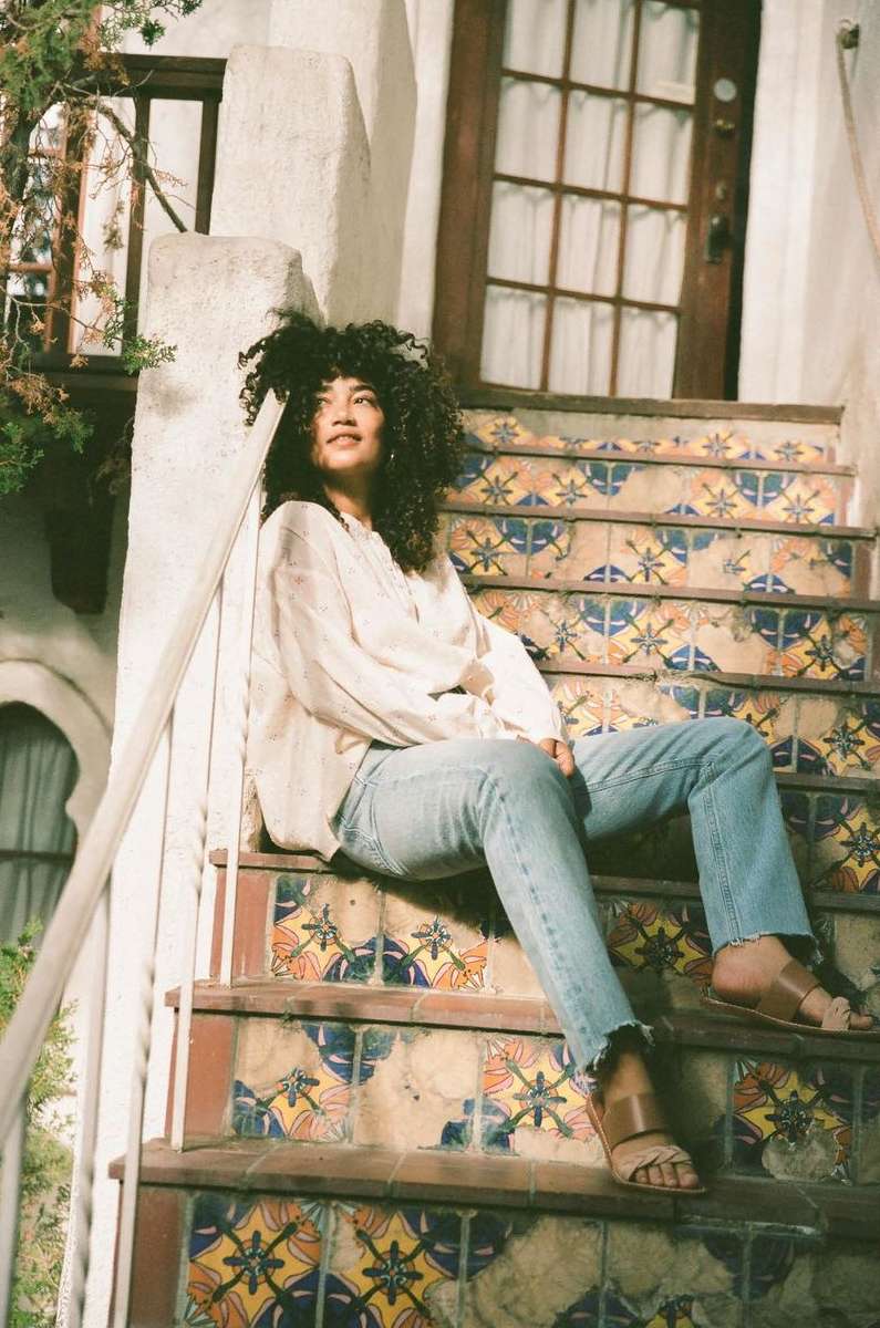 A woman with curly hair sits on patterned outdoor stairs in jeans and a loose blouse, enjoying the sunlight in a casual pose.