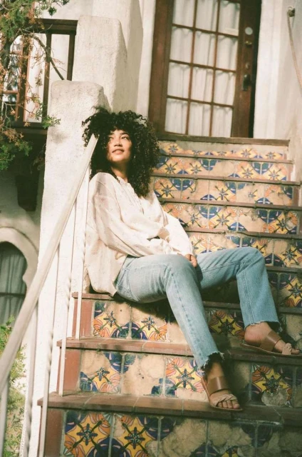 A woman with curly hair sits on patterned outdoor stairs in jeans and a loose blouse, enjoying the sunlight in a casual pose.
