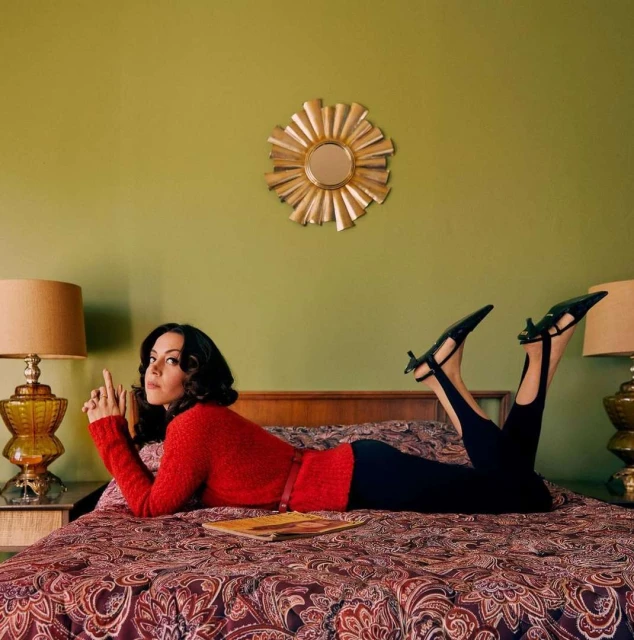 Aubrey Plaza is lying on a patterned bedspread in a red sweater and black pants in a retro-styled room with green walls and decor.
