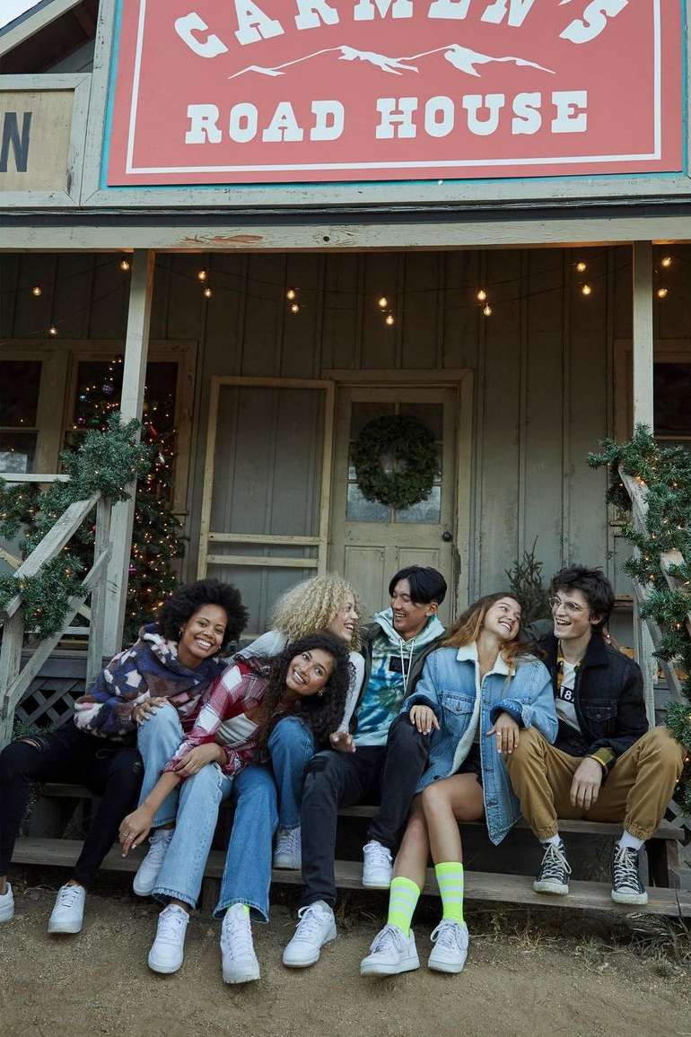 Group of six smiling young adults in casual clothing sitting on porch steps of Carmen's Road House decorated with holiday lights.