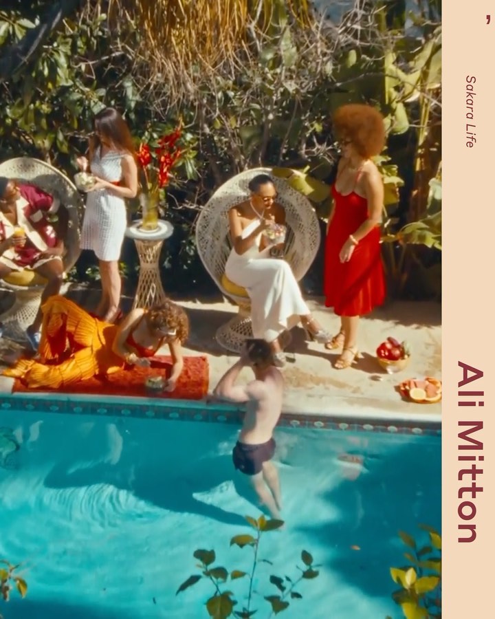 Group of people gather poolside for cocktails and conversation. Sunny outdoor setting. Women in dresses, man swimming.