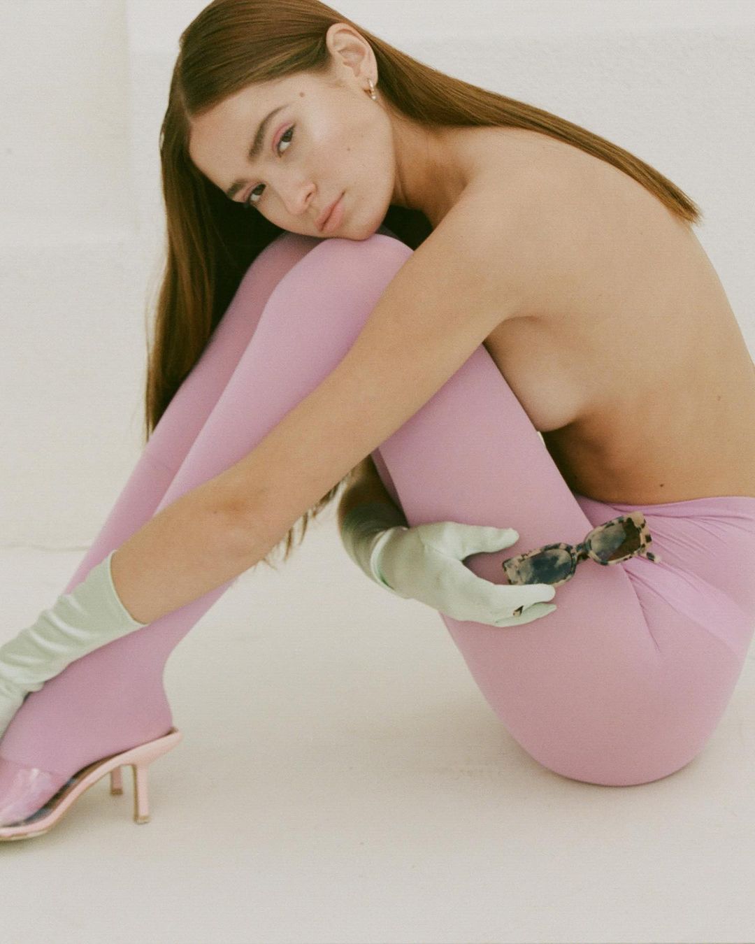A woman in pink leggings poses in a photo studio wearing light green gloves and pink high-heeled shoes and holding sunglasses.