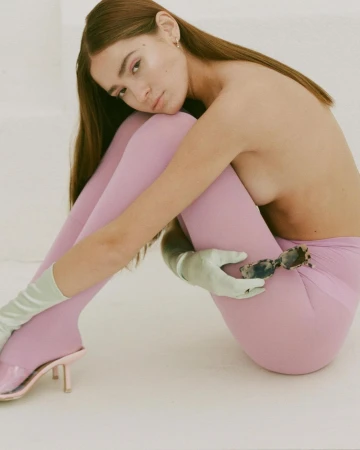 A woman in pink leggings poses in a photo studio wearing light green gloves and pink high-heeled shoes and holding sunglasses.