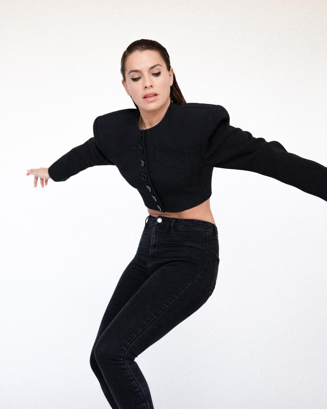 A woman in a black cropped jacket and black jeans poses in front of a white background, arms outstretched.