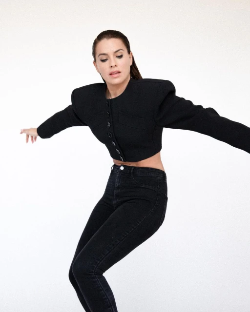 A woman in a black cropped jacket and black jeans poses in front of a white background, arms outstretched.