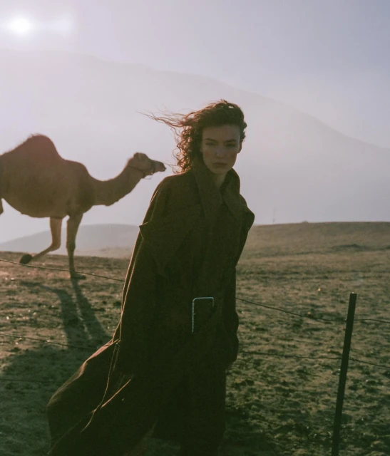 Fashion shot of a woman in a brown trench coat, posing outdoors with a camel, landscape, and mountains in the background.