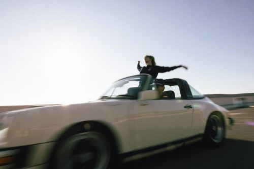 Two people in a vintage convertible car, one leaning out of the sunroof with arms raised against a blue sky, motion blur present.