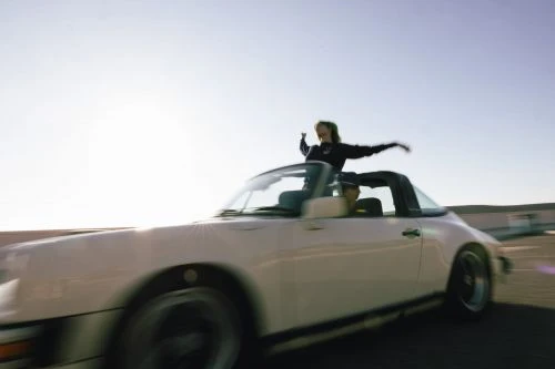 Two people in a vintage convertible car, one leaning out of the sunroof with arms raised against a blue sky, motion blur present.
