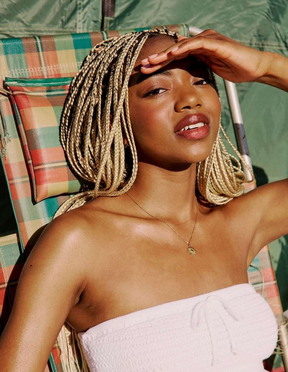 A woman with blonde braids reclines in a lawn chair with a plaid pattern, shading her eyes with one hand in outdoor lighting.