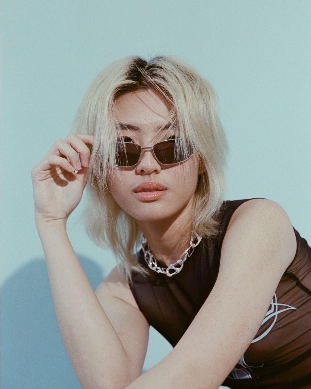Portrait of a young person with bleached blonde hair wearing sunglasses and a dark mesh top, by photographer Ali Mitton.