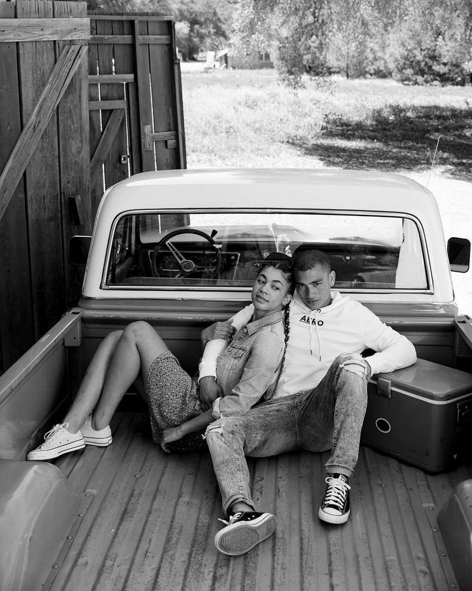 Monochromatic shot of a couple sitting in the back of a pickup truck in a rural setting, wearing jeans and sneakers, with a cooler nearby.