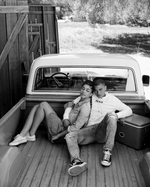 Monochromatic shot of a couple sitting in the back of a pickup truck in a rural setting, wearing jeans and sneakers, with a cooler nearby.