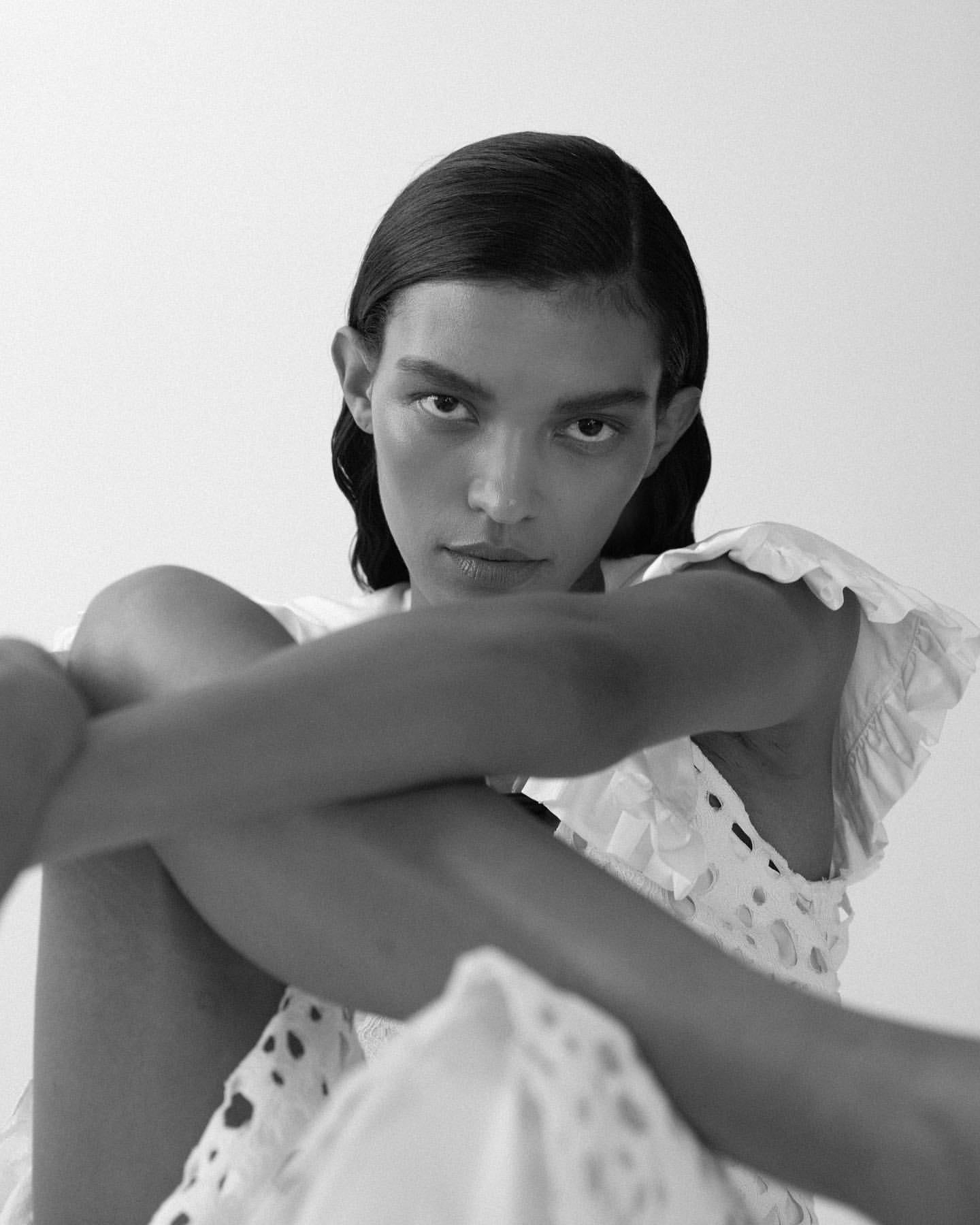 Black and white portrait of a woman with wet hair in a white, eyelet dress, posing against a neutral background in a studio.