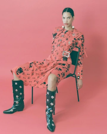 Model seated on a black chair wearing a pink floral dress and black knee-high boots against a pink background