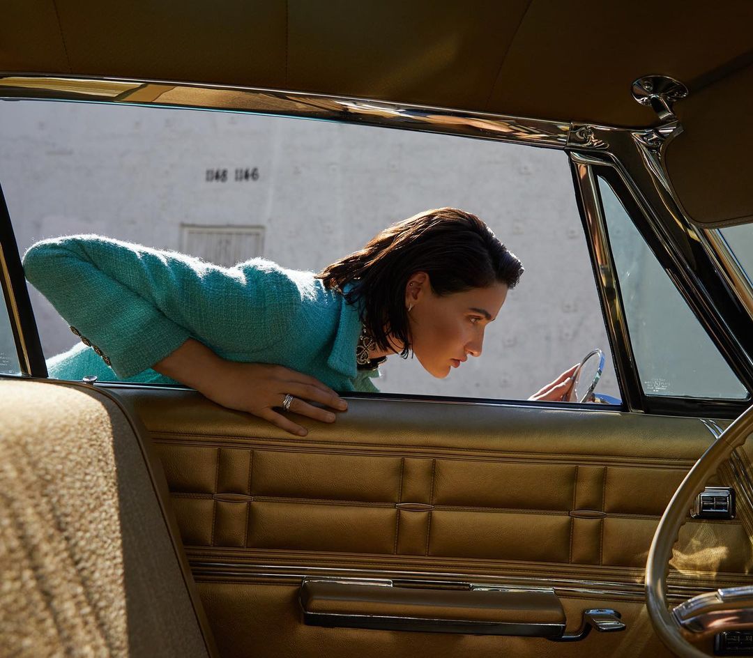 A woman peers out of a gold car window in a turquoise jacket, accented by the building's neutral backdrop and sunlit setting.