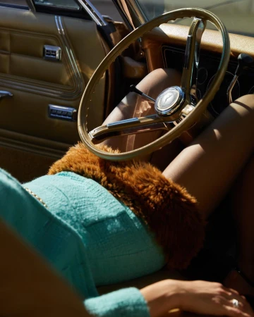 Woman in a blue dress sitting inside a vintage car with a tan interior, focusing on the steering wheel and the car's design.