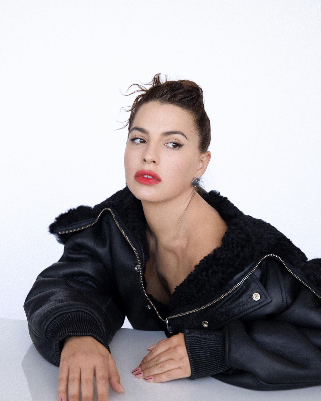 Brunette woman with red lipstick and a black leather jacket looks off camera in a medium shot portrait against a bright white background.