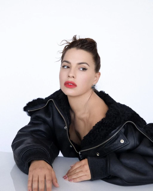 Brunette woman with red lipstick and a black leather jacket looks off camera in a medium shot portrait against a bright white background.