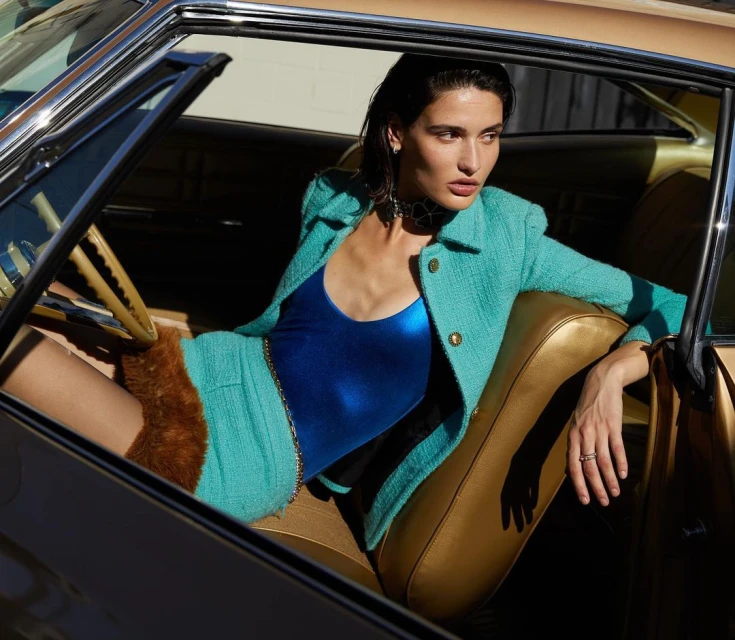 A woman in a blue bodysuit and turquoise tweed suit sits inside a classic car with gold interior and dark window frames, fashion shoot.